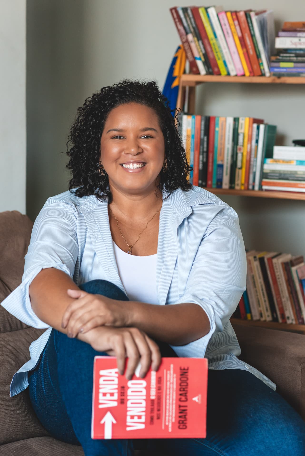 Karla Manu sentada em um sofá segurando o livro Venda ou Vendido